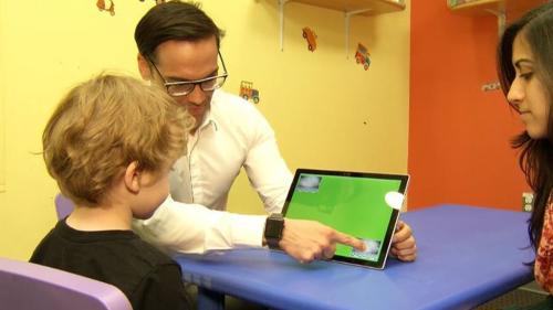 A man with dark hair and glasses, Dr. Francys Subiaul, sits at a table in a colorful room and shows a child a screen while a woman sits with them and observes.